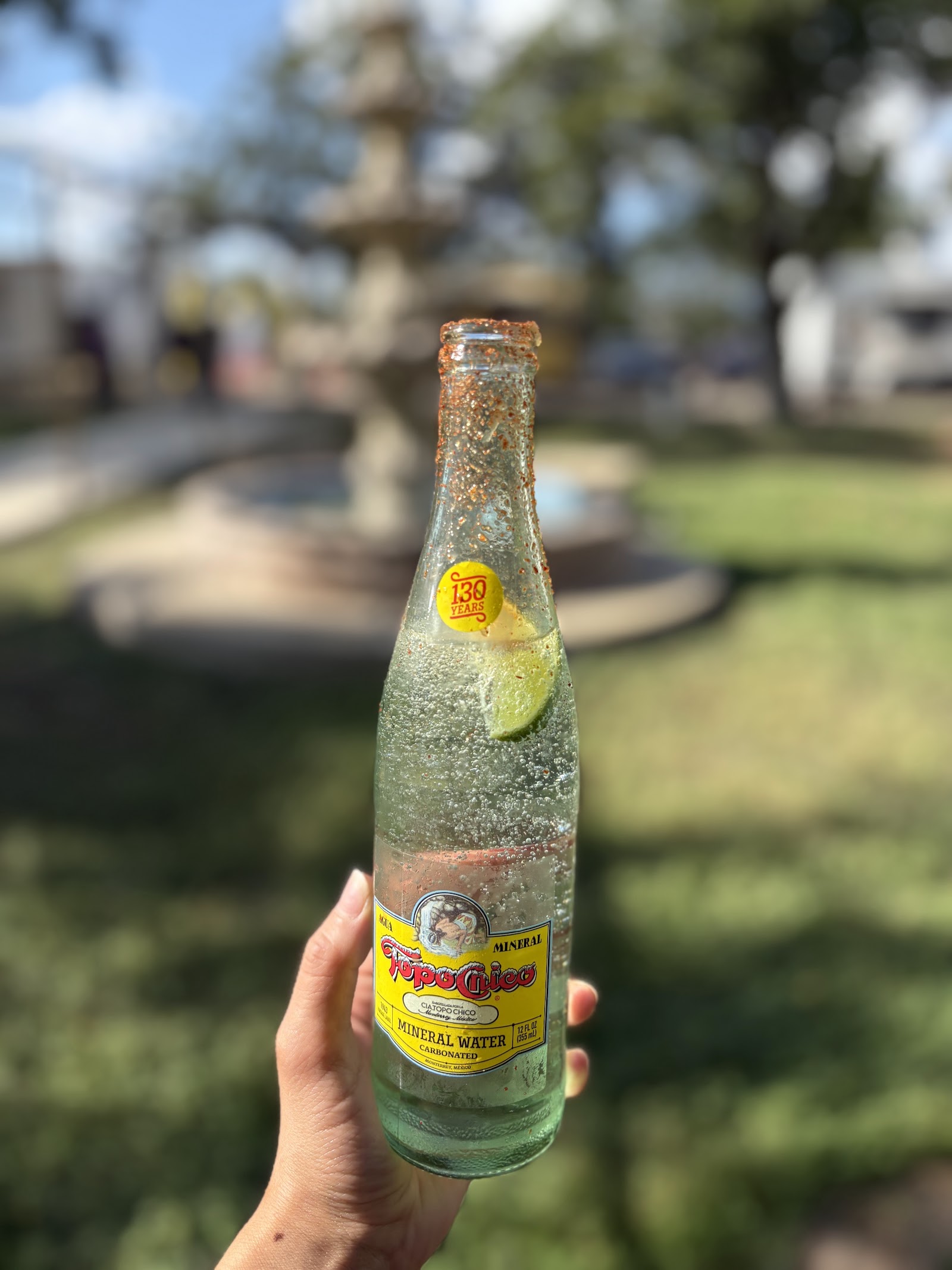 Chili-lime rimmed Topo Chico mineral water held up in front of the stone fountain
